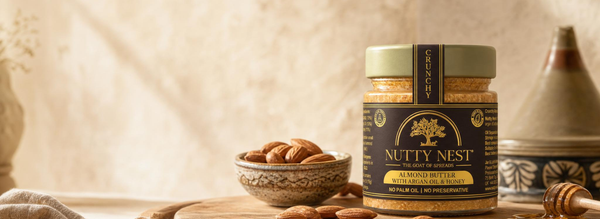 A jar of Nutty Nest Almond Butter stands on a wooden surface with scattered almonds, a small bowl of almonds, and a rustic background. The jar has a dark brown label with gold lettering and a light olive-green lid.